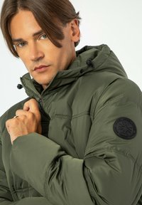 Jasnozielona kurtka puchowa z zamkiem błyskawicznym, regulowanym kapturem i czarną okrągłą naszywką na rękawie. Gładka tekstura i pikowany wzór.