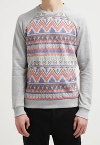 Sudadera gris con un patrón geométrico multicolor que incluye flechas, triángulos y acentos circulares en la parte delantera, y mangas raglán.