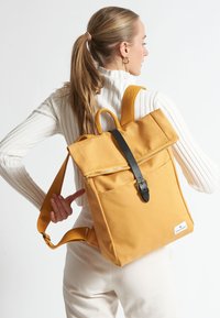 Zaino in tela gialla con design a risvolto, chiusura con cinturino in pelle nera, tasca frontale con cerniera e spallacci in tessuto regolabili.