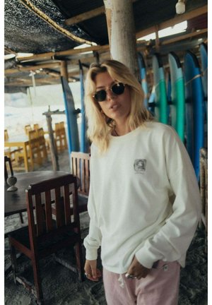 Osoba o blond włosach, w okularach przeciwsłonecznych, białej bluzie i różowych spodniach stoi w pomieszczeniu obok desek surfingowych i drewnianych krzeseł w zacienionej okolicy przy plaży.