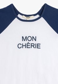 Hvid T-shirt med marineblå raglanærmer, med teksten "MON CHÉRIE" i fed marineblå skrift. Blødt bomuldsstof, rund halsudskæring.