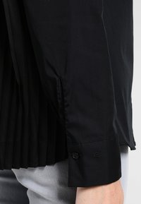 Black long-sleeve shirt featuring a pleated back, buttoned cuffs, and subtle embroidered detail near the cuff, made from smooth fabric.