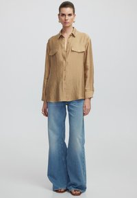 Beige strukturierte Knopfhemd mit zwei Brusttaschen, kombiniert mit hellblauen Schlagjeans. Glattes Material, entspannter Schnitt und lässiges Design.