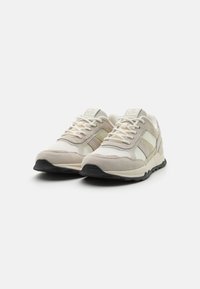 Zapatillas beige con una mezcla de materiales de ante y malla, acentos claros, cordones blancos y una suela de goma negra. Diseño atlético clásico.