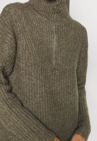 Pull tricoté vert olive, doté d'un col montant, d'une demi-fermeture éclair à l'avant et d'une finition texturée avec des manches côtelées.