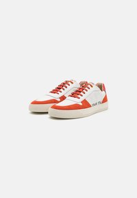 Hvite sneakers med røde semskede detaljer, med rund tå, kontrastfargede snørebånd og en teksturert gummisåle. Merking synlig på siden.