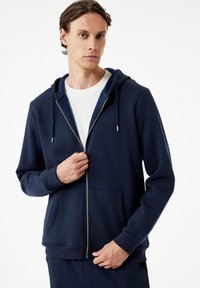 Joven con una sudadera azul marino con cremallera sobre una camisa blanca, sosteniendo la cremallera con una mano, frente a un fondo blanco plano.