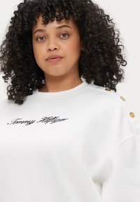 Hvid sweatshirt med broderet "Tommy Hilfiger" logo, der har guldknaffekanter på skulderen og et afslappet fit design.