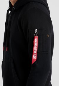 Felpa nera con cappuccio e tasca con zip sulla manica sinistra, caratterizzata da un'etichetta rossa "RIMUOVERE PRIMA DEL VOLO". Tessuto morbido e cordini regolabili.