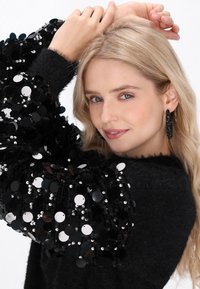 Črna pulover s oversize črnimi bleščečimi rokavi. Model ima svetlo blond lase. Uhani so veliki in črni, teksturirani z zapletenimi detajli.