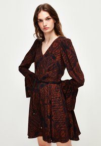 Braun gemustertes Wickelkleid mit langen Glockenärmeln, tiefem V-Ausschnitt und tailliertem Schnitt, der durch einen schwarzen Bindegürtel betont wird. Leichtes Material.