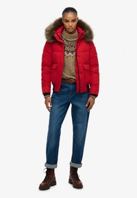 Rød puffjakke med pelsforet hætte, parret med en tan strikket sweater og blå oprullede jeans. Brune snørebåndsstøvler fuldender looket.