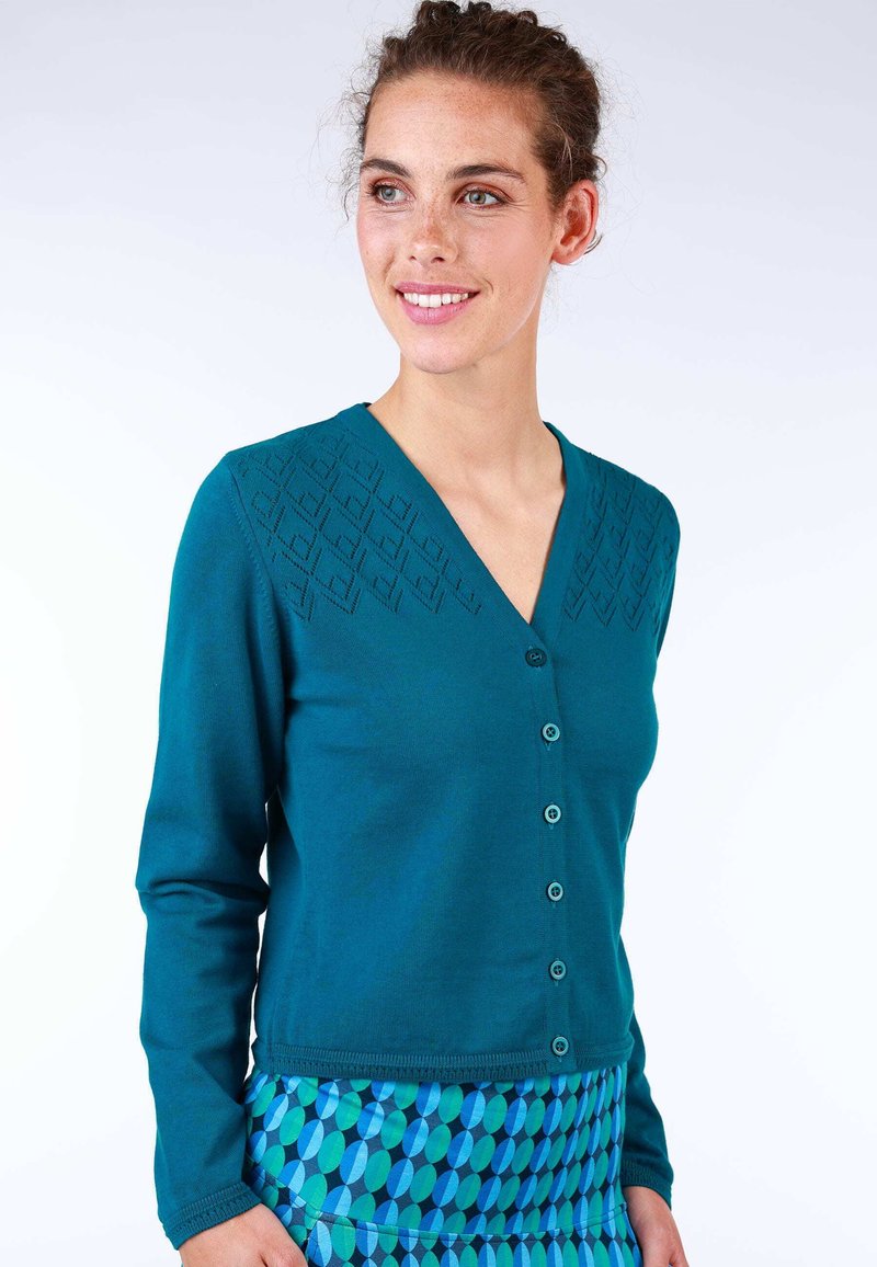 Türkiser Cardigan mit langen Ärmeln, V-Ausschnitt, strukturiertem Muster an den Schultern und sechs Knöpfen. Über einem gemusterten blauen Rock getragen.