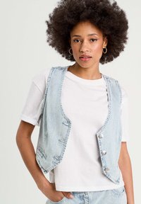 Gilet en denim à rayures avec une coupe décontractée, doté de cinq boutons et de poches arrière. Porté sur un t-shirt blanc uni.