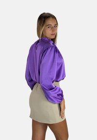 Lila Satinbluse mit schmalen Ärmel-Puffärmeln und Rückenschlitz-Detail, kombiniert mit einem taillierten beigen Rock, der glatte Texturen und eine moderne Silhouette präsentiert.
