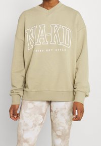 Sudadera en color caqui con escote en V, corte oversized y texto gráfico blanco "NA-KD NOTHING BUT STYLE" en el frente. Puños y dobladillo acanalados.