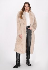 Lange beige nepbontjas met een reverskraag; zachte textuur, open voorkant. Gekozen boven een witte top en donkerdenim, aangevuld met zwarte kniehoge laarzen.