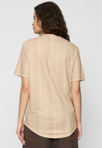Beige Kurzarmoberteil mit vertikalen weißen und pastellfarbenen Streifen, abgerundetem Saum und glatter Textur. Keine sichtbaren Logos oder Verzierungen.