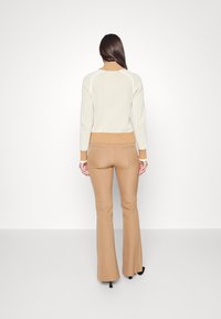 Kobieta stojąca tyłem, ubrana w kremowy i tanowy prążkowany sweter oraz tanowe spódnice w kształcie dzwonu, na czarnych szpilkach, na białym tle.