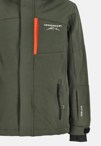 Olivgrön väderbeständig jacka med en orange detalj, ficka med dragkedja och logotyp. Har justerbara manschetter och en slät yta.