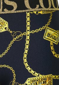 Tajts med svart bas som har ett glänsande mönster av gyllene kedjor och etiketter, som framhäver en djärv logotyp vid midjan i glittrande guld.