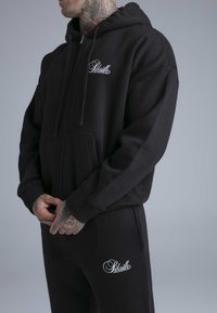 Zwarte hooded trui met voorzakken en logo op de linkerborst, gecombineerd met zwarte joggingbroek met logo op de rechterdij. Zacht materiaal.