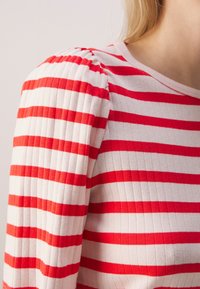 Gros plan d'un haut en tissu côtelé blanc avec des rayures rouges horizontales et un détail bouffant à l'épaule.