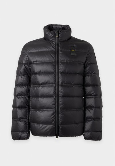 Veste d'hiver noire matelassée avec quilting horizontal, col montant, fermeture éclair à l'avant et un petit logo jaune et noir sur la poitrine.