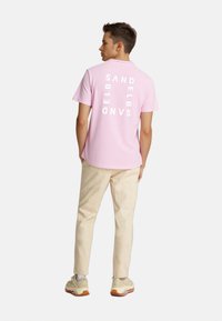 Pink t-shirt with vertical white text on the back. Model wears cream-colored pants and beige shoes. Smooth cotton fabric, casual fit.