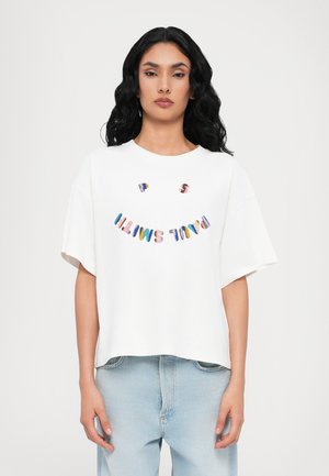 T-shirt blanc surdimensionné à manches courtes, avec un texte irrégulier multicolore épelant "P S SMILE" dans une formation souriante.