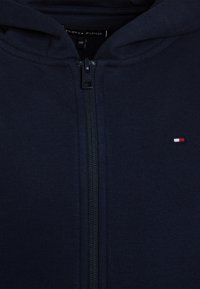 Mörkblå zip-up huvtröja i mjukt tyg, med dragkedja framtill och en liten broderad logotyp i röd, vit och blå på vänster bröst.