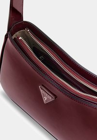 Burgundowa syntetyczna torebka na ramię o połyskującej powierzchni, z zamykanymi na suwak kieszeniami i metalowym logotypem z przodu. Wnętrze wykończone jest tkaniną w kontrastowym kolorze.
