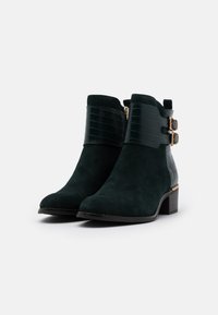 Botines de ante verde que presentan un panel de textura de cocodrilo, hebillas en tono dorado y un tacón grueso.