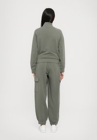 Vrouw met lang donker haar, gekleed in olijfgroene cargojoggingbroek en bijpassend zip-up jack, met haar rug naar de camera op een witte achtergrond.