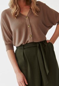 Brązowy sweter z dzianiny z dekoltem w serek, krótkimi rękawami i trzema guzikami. Połączony z zielonymi spodniami o wysokim stanie z przewiązanym paskiem.