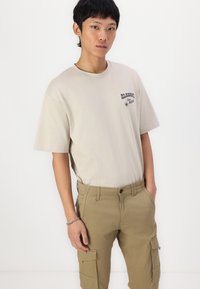 Beige katoenen T-shirt met een ronde hals, korte mouwen en bedrukte tekst; gecombineerd met khaki cargobroek met zijzakken.
