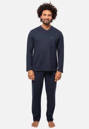 Uomo in piedi a piedi nudi che indossa una camicia a righe blu scuro a maniche lunghe e pantaloni tinta unita abbinati su uno sfondo chiaro e uniforme.