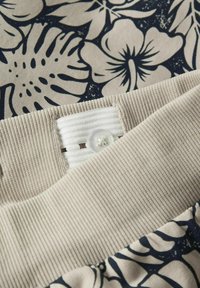 Ceinture beige avec un tissu côtelé texturé, ornée d'un bouton blanc et d'une boucle en tissu. L'arrière-plan présente un motif floral marine avec de grandes fleurs.