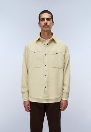 Uomo in piedi rivolto in avanti che indossa una camicia beige in velluto a coste abbottonata sopra una felpa crema e pantaloni marrone scuro su uno sfondo chiaro uniforme.