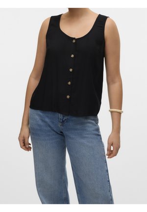 Vrouw die een mouwloze zwarte blouse met knopen draagt, gecombineerd met lichtblauwe spijkerbroek en een beige armband aan de linkerpols.