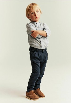 Jeune garçon aux cheveux blonds debout les bras croisés, portant une chemise gris clair à boutons, un pantalon sombre et des chaussures en daim marron sur un fond uni.