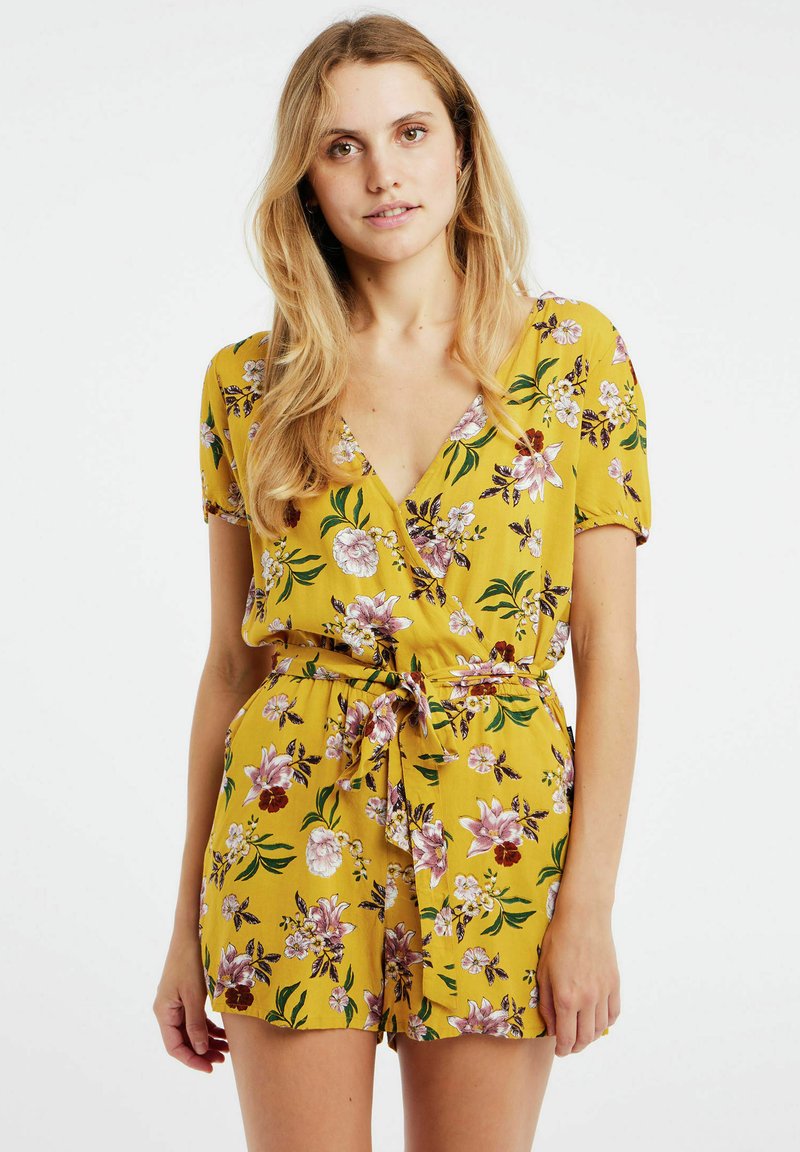 Combinaison florale jaune à manches courtes, décolletée en V et taille ceinturée. Présente un motif de fleurs violettes et blanches sur un tissu texturé.