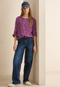 Blusa viola a fantasia con apertura a occhiello, abbinata a jeans larghi blu scuro e sneaker bianche. La modella indossa un cappellino da baseball blu.