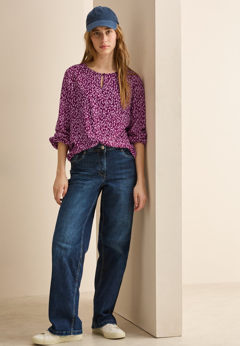 Blusa viola a fantasia con apertura a occhiello, abbinata a jeans larghi blu scuro e sneaker bianche. La modella indossa un cappellino da baseball blu.