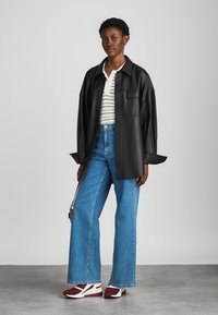 Chemise surdimensionnée en cuir noir, chemise rayée blanche et noire en dessous, jeans larges bleus et baskets bordeaux et blanches.