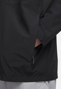 Μαύρο αδιάβροχο μπουφάν με μπροστινή τσέπη, ρυθμιζόμενο κορδόνι στο τελείωμα και λεία υφή υφάσματος.