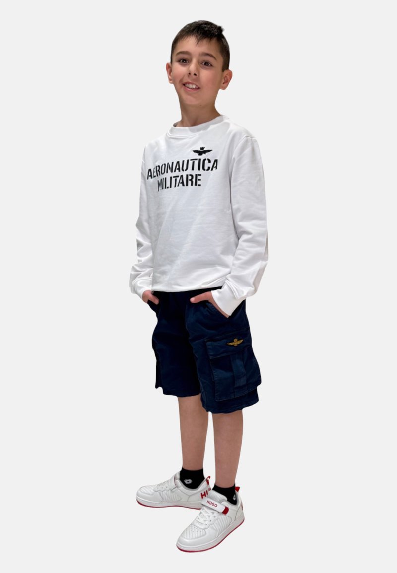 Camicia bianca a maniche lunghe con testo nero "Aeronautica Militare", pantaloni cargo blu navy con tasche e sneakers bianche con dettagli rossi.