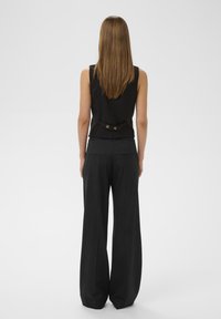 Black sleeveless blouse with a fitted design and bow detail at the back, paired with loose-fitting black trousers featuring side pockets.