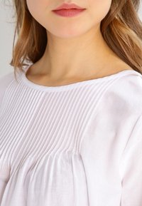 Hvid bluse med vertikale pintuck-detaljer på brystet og blødt, let tekstureret stof, med rund halsudskæring og sammensnørede ærmer.
