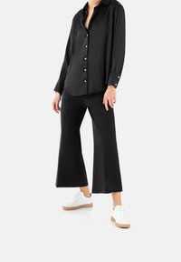 Camicia in satin nera a maniche lunghe con bottoni, abbinata a pantaloni neri a gamba larga e sneakers bianche con suola beige.